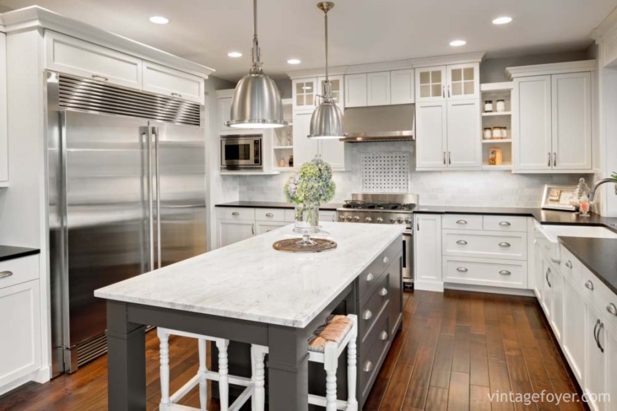 High end stainless steel appliances, white marble countertop on a grey island, custom white shaker cabinetry with a black quartz countertop, and a dark toned hardwood flooring. 